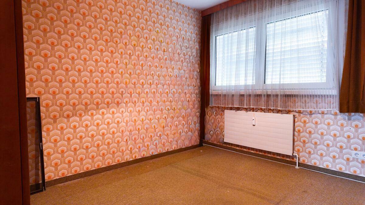 Zimmer mit auffälliger gemusterter Tapete und einem Heizkörper unter dem Fenster.