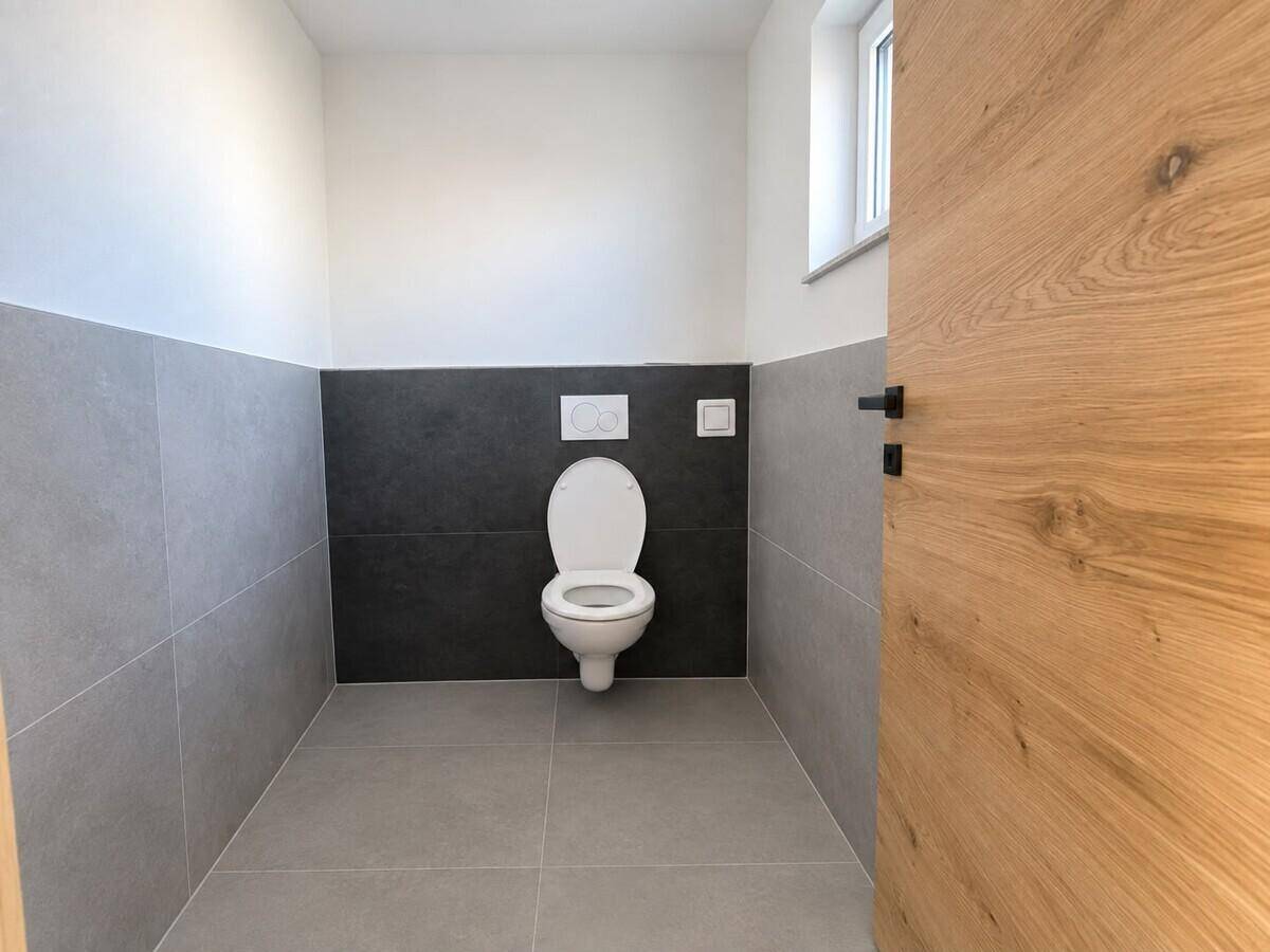 Modernes Gäste-WC mit wandhängender Toilette, grauen Fliesen und einem kleinen Fenster für Tageslicht.