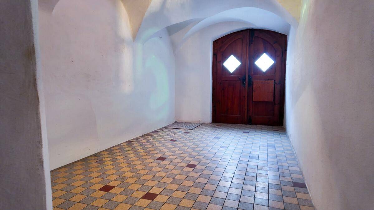 Historischer Eingangsbereich mit Gewölbedecke und gemusterten Fliesen, führt zu einer Holztür.