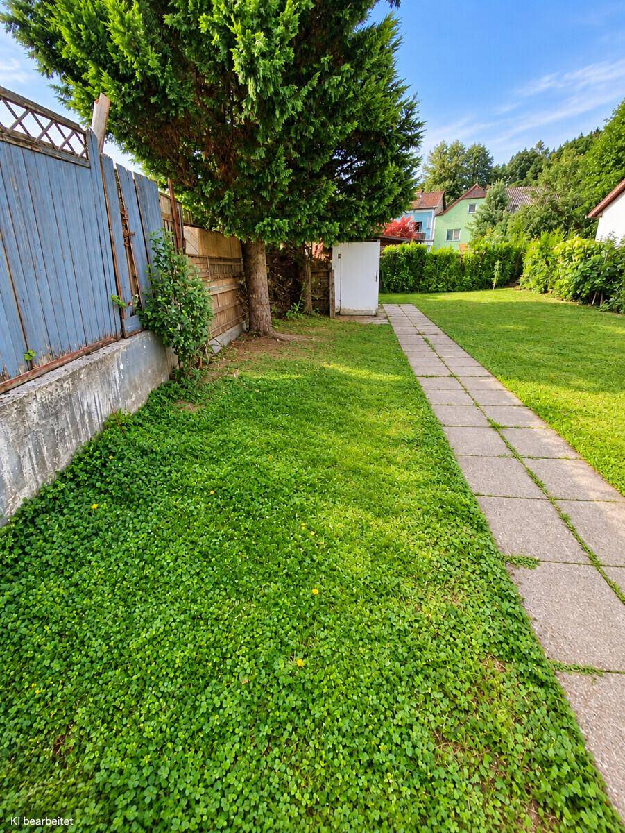 Grüner Gartenbereich mit Rasenfläche, einem gepflasterten Weg und hohen Bäumen.