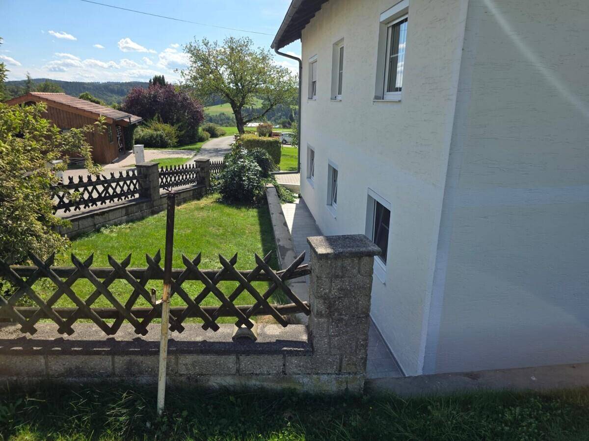 Seitenansicht eines weiß verputzten Hauses mit einem kleinen, eingezäunten Gartenbereich unter strahlendem Sonnenschein.
