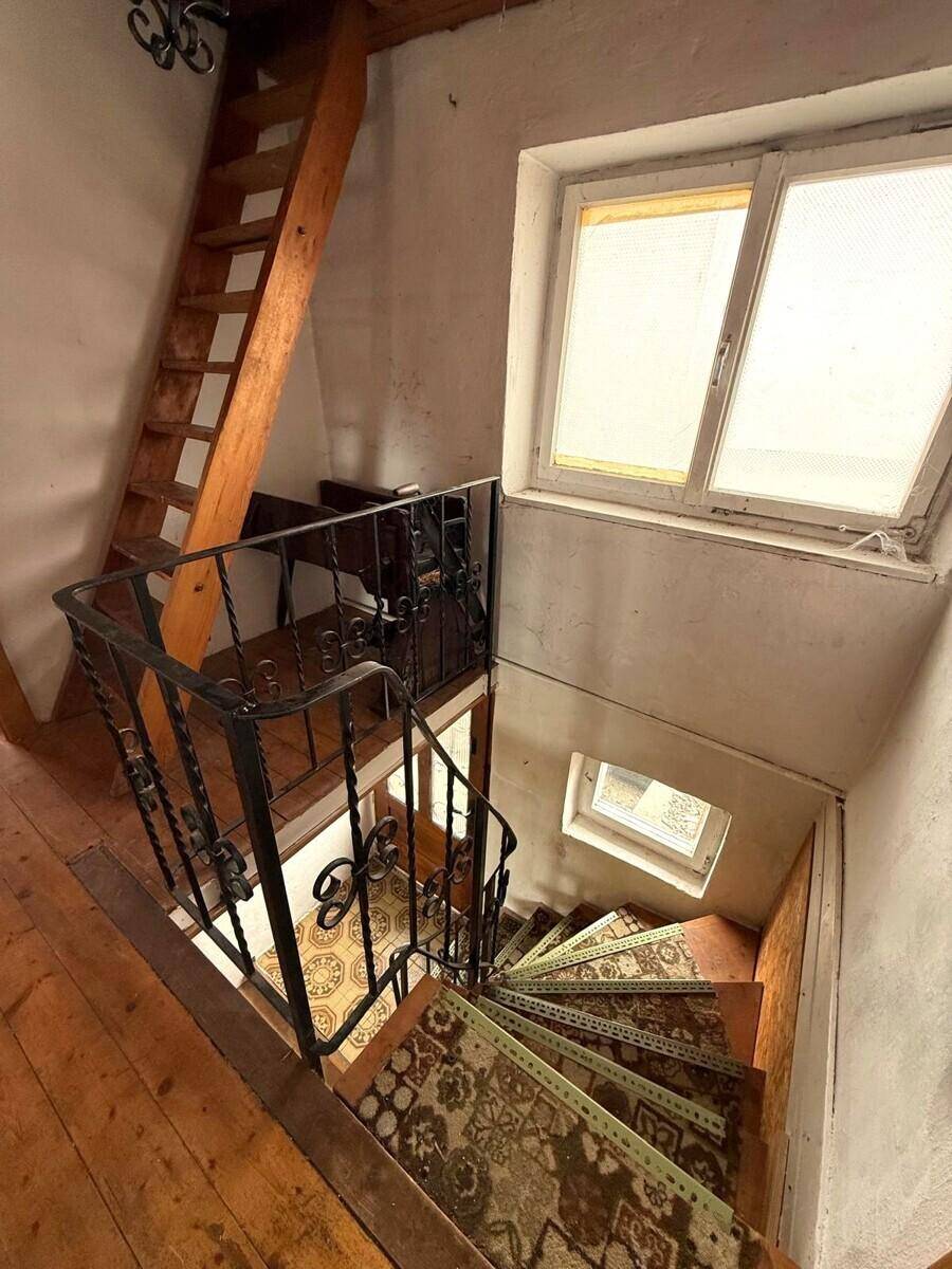 Historische Holztreppe mit schmiedeeisernem Geländer und Teppichläufer, beleuchtet durch ein Fenster.