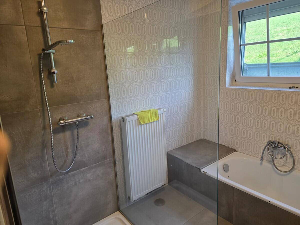 Badezimmer mit Duschkabine, Badewanne, grauen Fliesen und einem Fenster für Tageslicht.