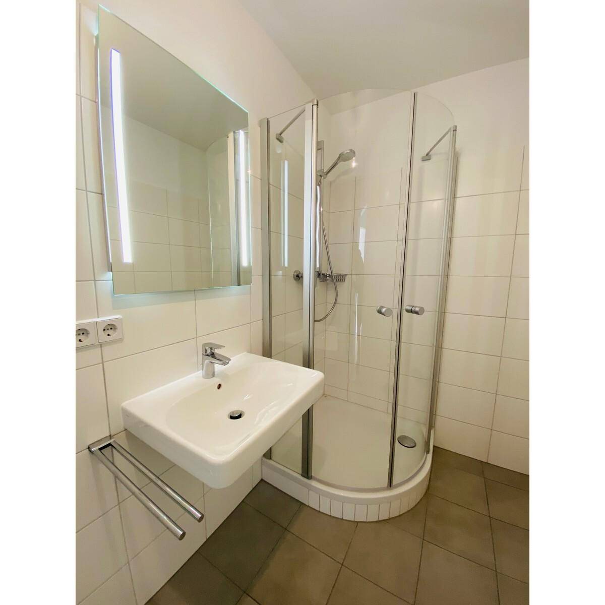 Modernes Badezimmer mit Duschkabine, Einzelwaschbecken und beleuchtetem Spiegel.