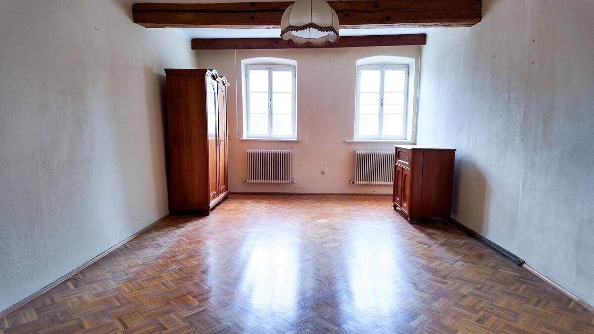 Heller Raum mit Holzbalken an der Decke, Parkettboden und zwei Fenstern.