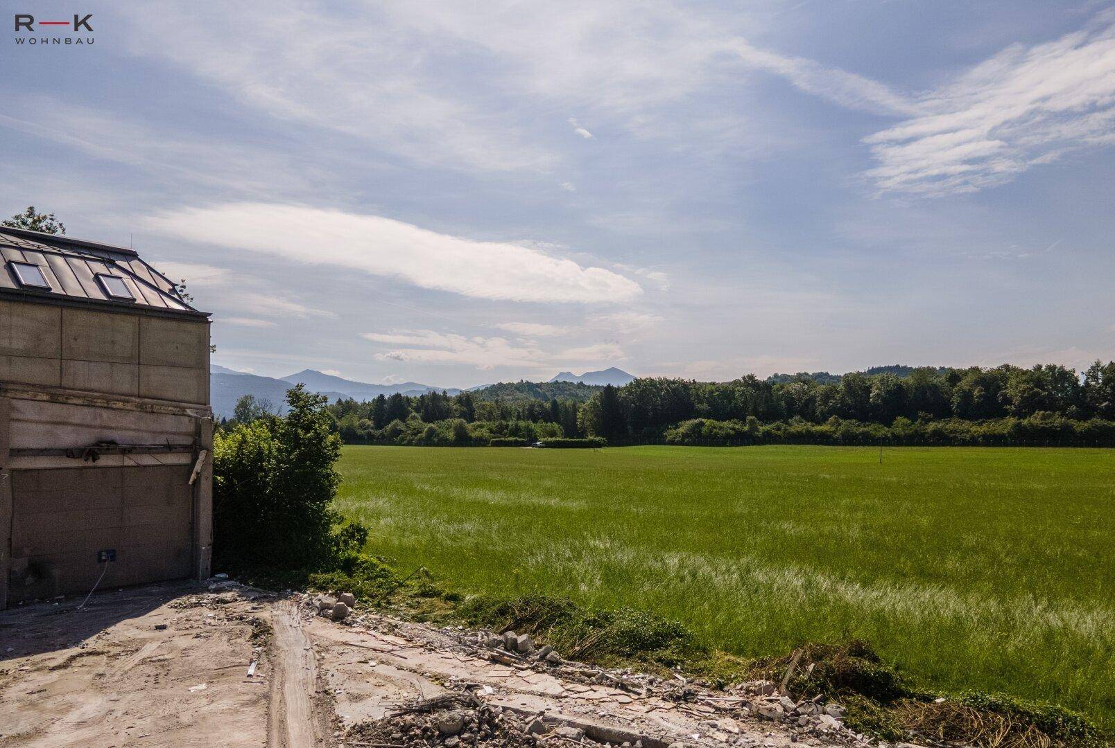 Blick über grüne Felder und Wälder zu den Bergen, mit einem teilweise abgerissenen Gebäude im Vordergrund.