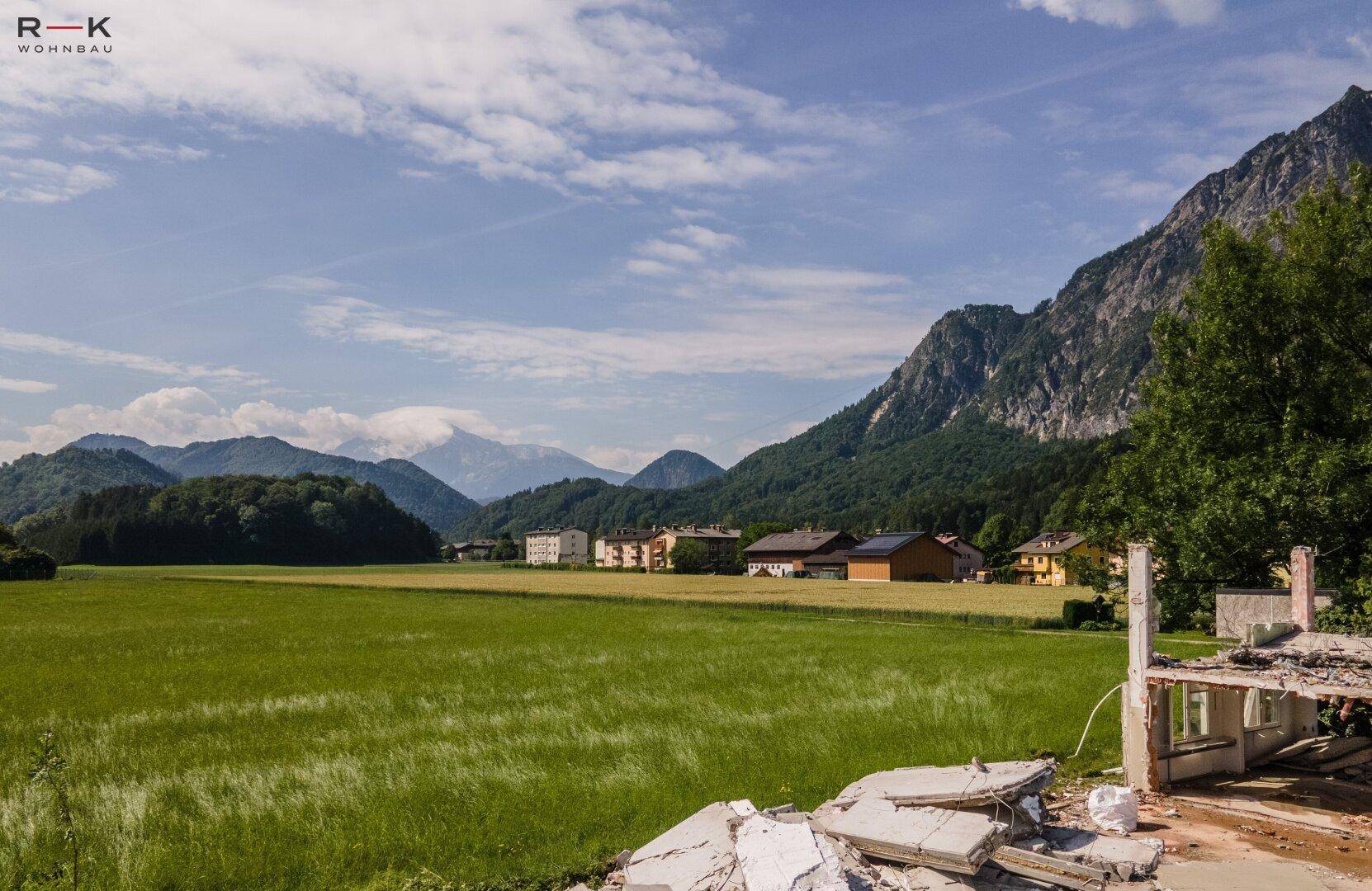 Panoramablick auf grüne Felder und majestätische Berge, mit Gebäuden in der Ferne und einer Baustelle im Vordergrund.