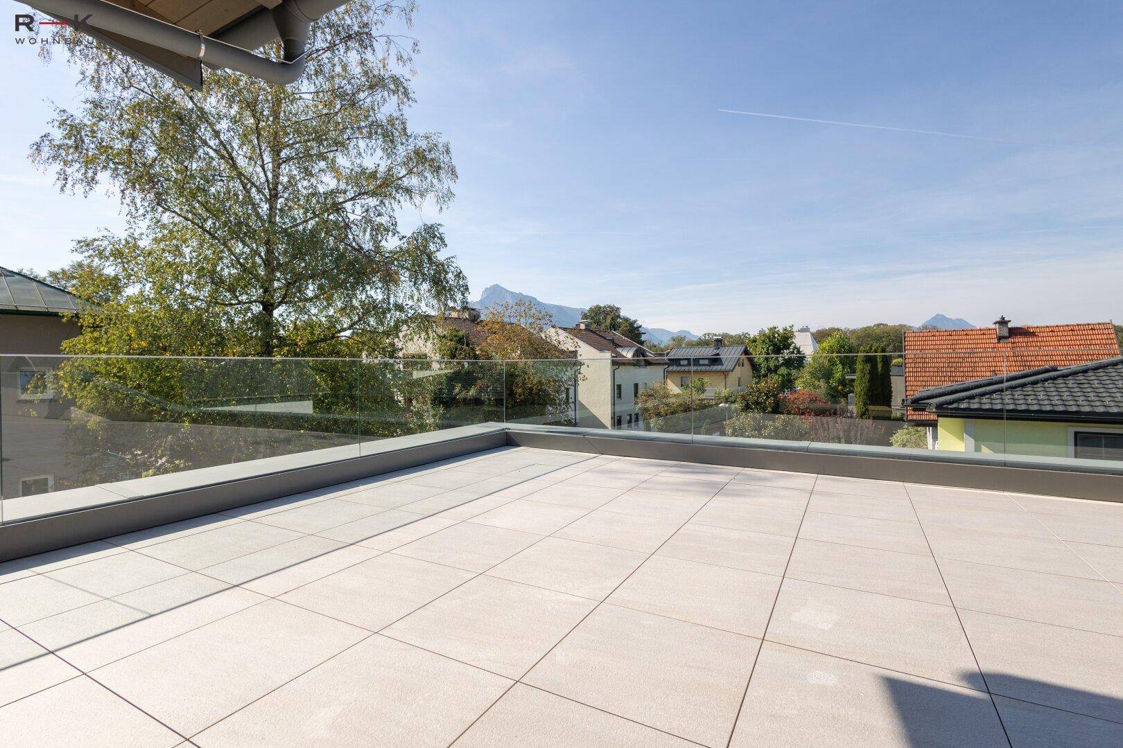 Große Dachterrasse mit modernen Fliesen und Glasbrüstung, umgeben von grüner Landschaft und Bergen.