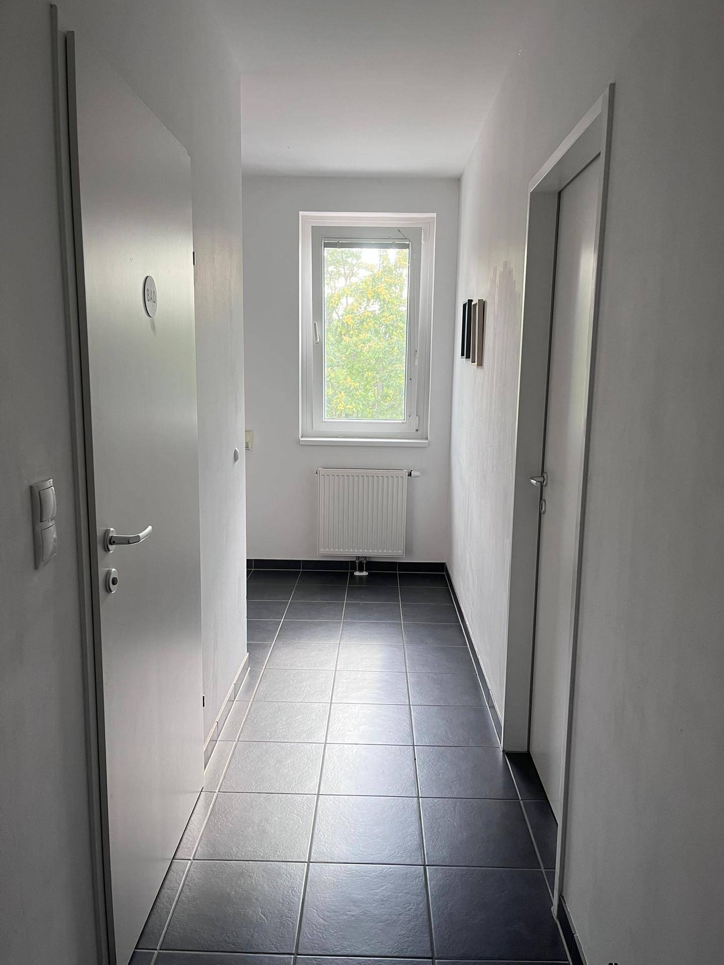 Heller Flur mit dunklen Fliesen und einem Fenster am Ende, das Tageslicht spendet.