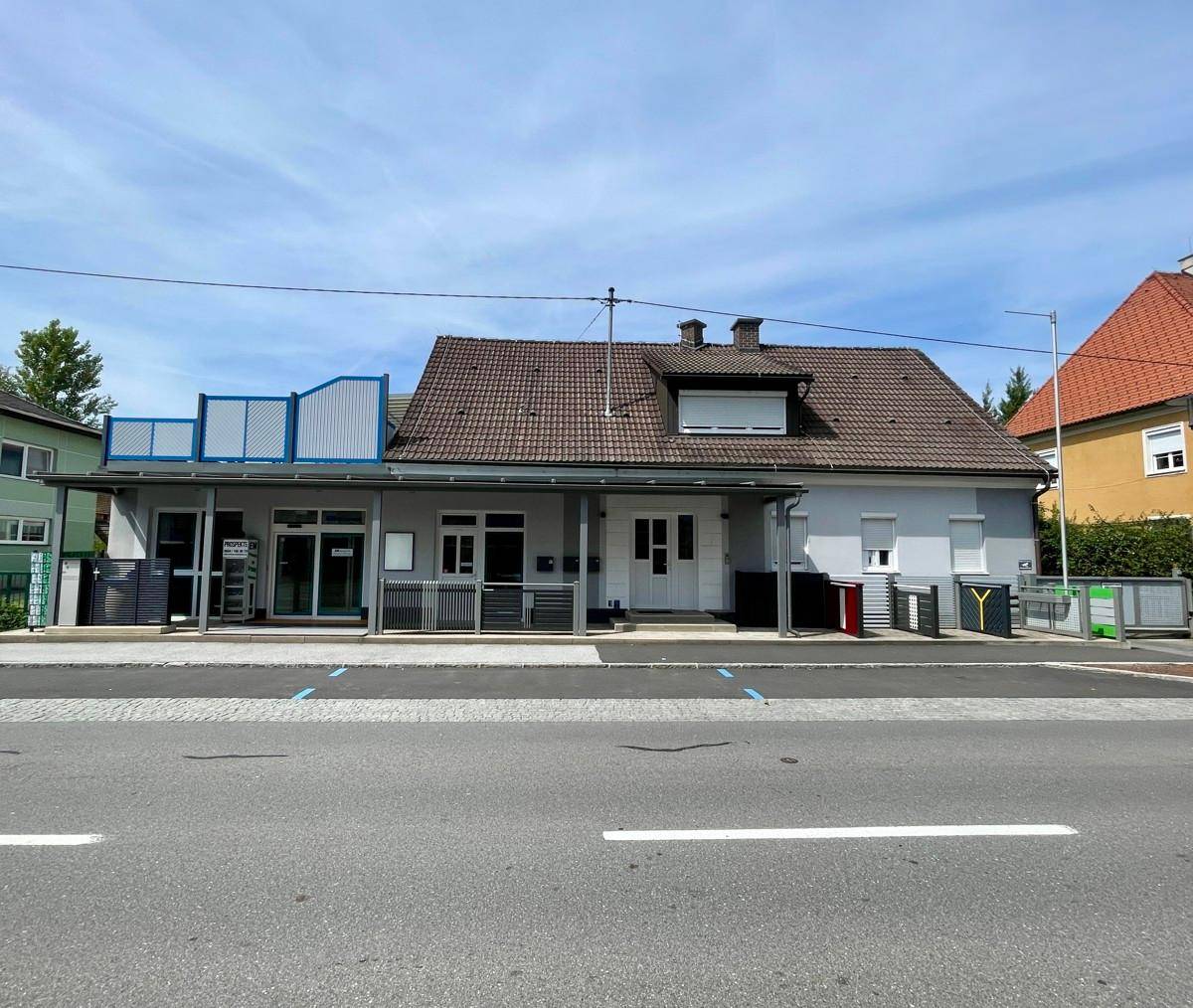 Vorderansicht des Gebäudes mit überdachtem Eingang, Rollläden und Parkplätzen an der Straße.