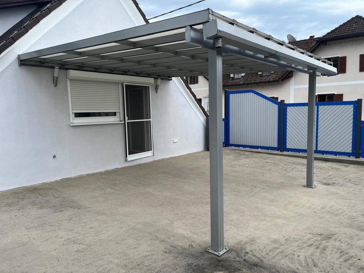 Überdachter Stellplatz mit Metallkonstruktion und Rollladenfenster am angrenzenden Gebäude.