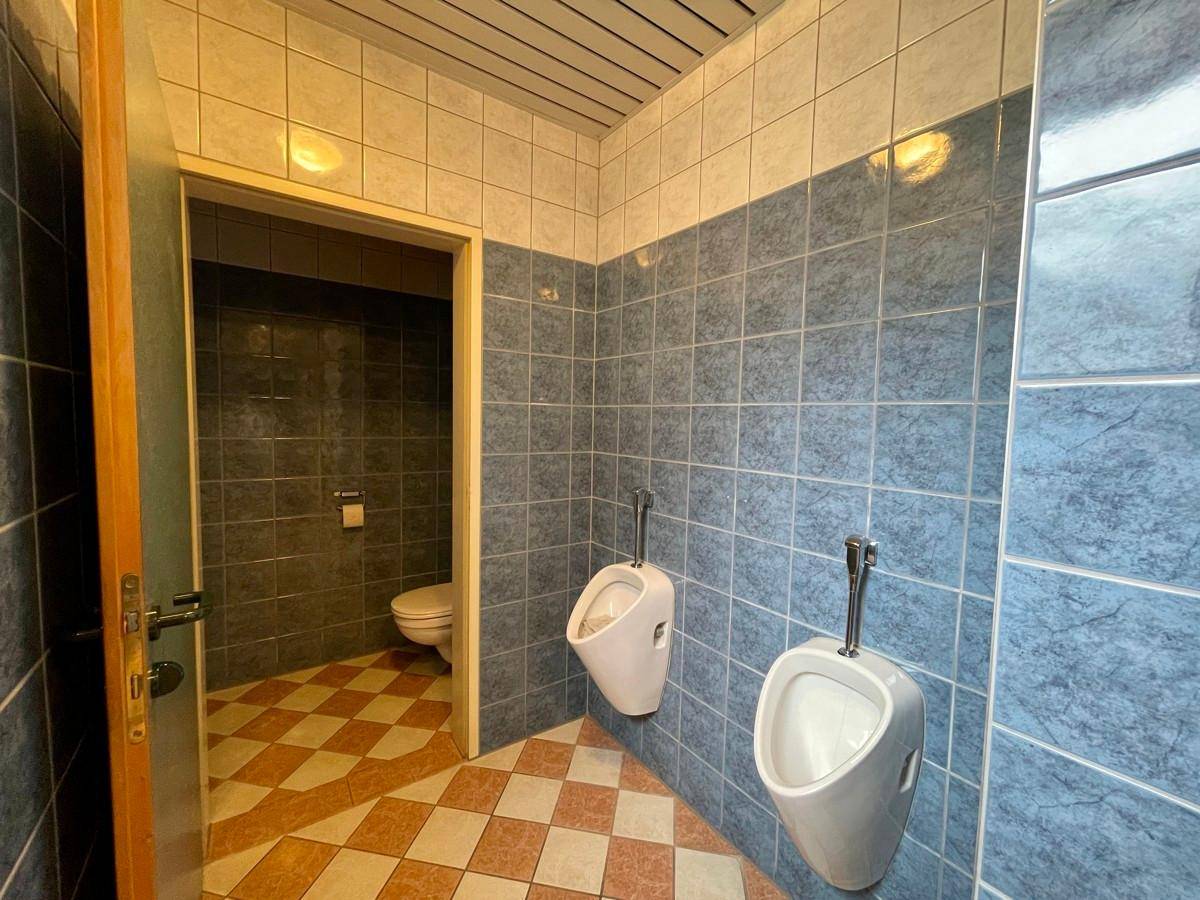 Gefliester WC-Bereich mit Urinalen und Zugang zu weiteren Toilettenkabinen.
