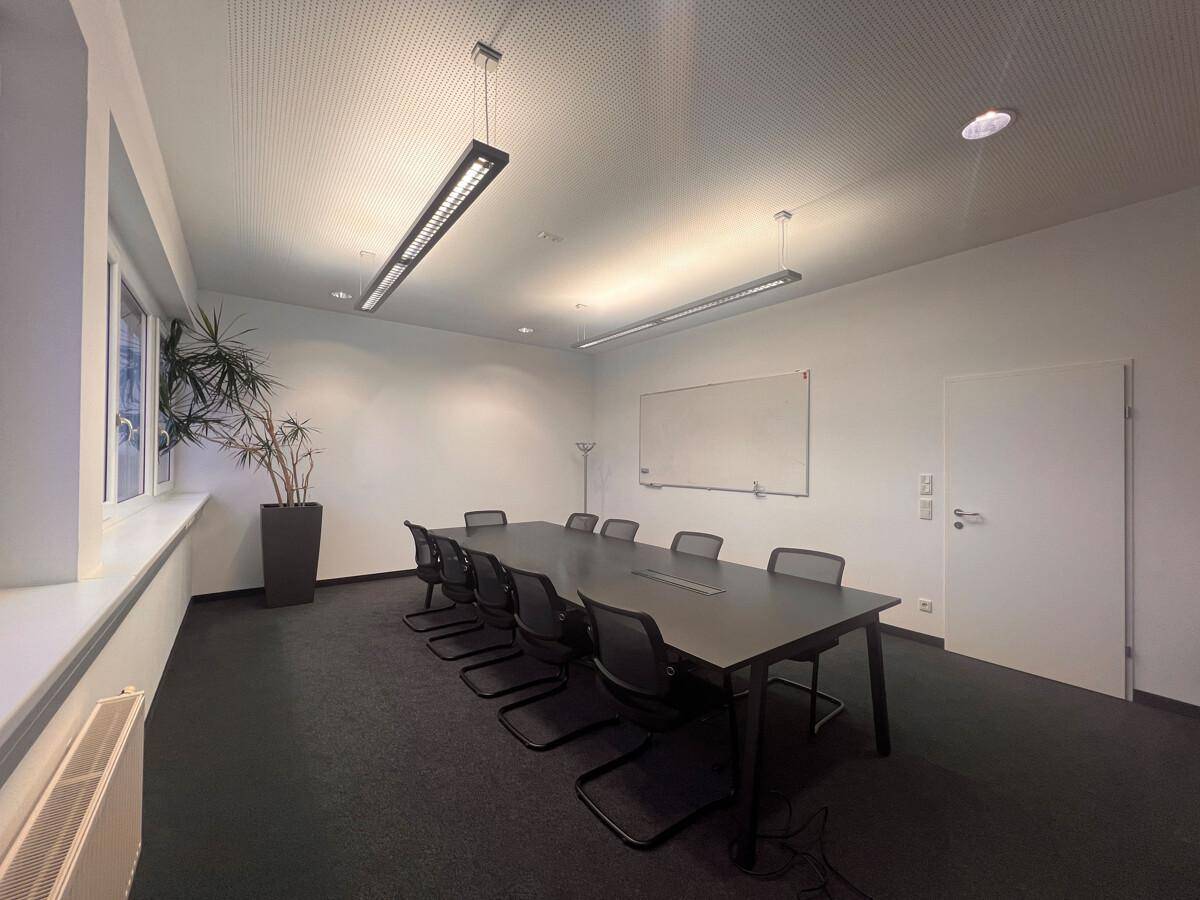 Konferenzraum mit großem Tisch, Stühlen und einem Fenster, das Tageslicht hereinlässt, ideal für Besprechungen.