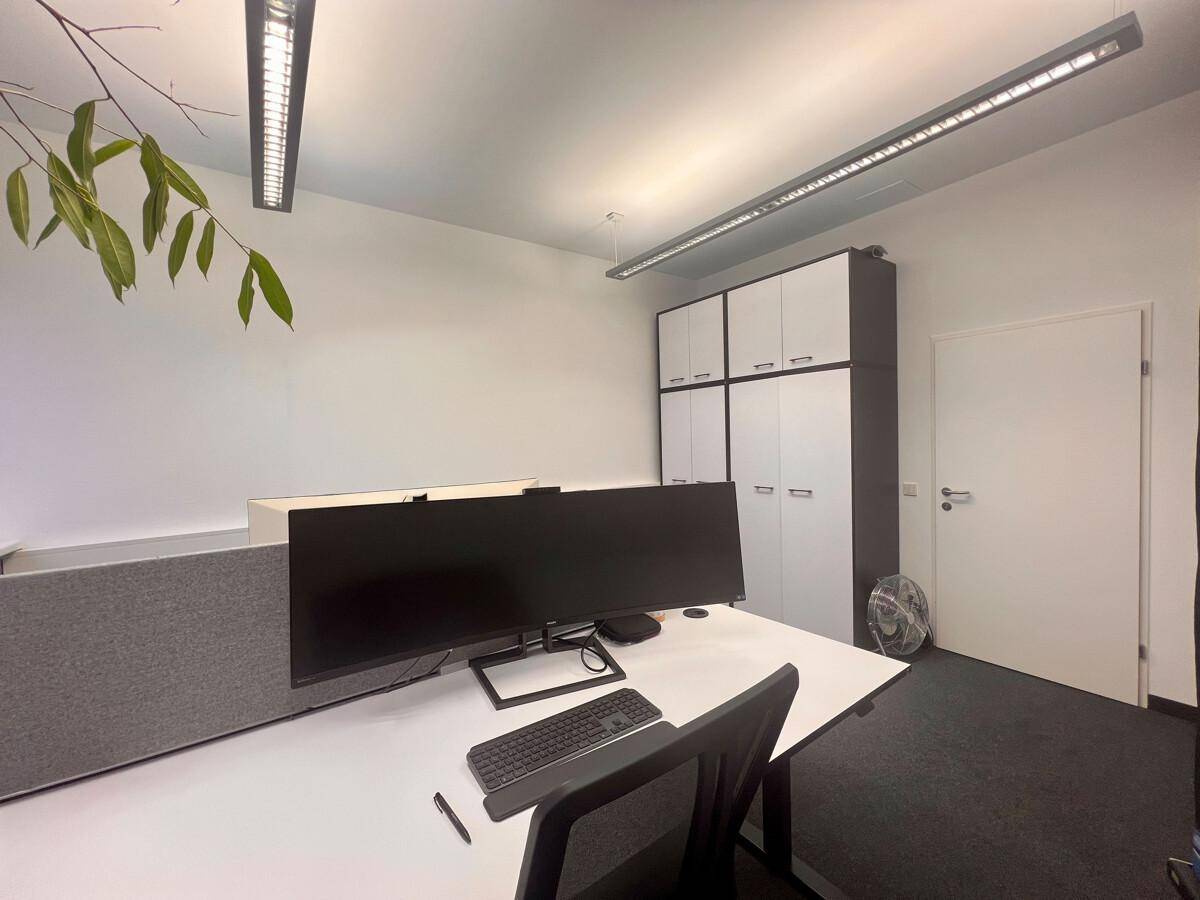 Heller Büroraum mit Arbeitsplatz, Schreibtisch und ergonomischem Stuhl, ergänzt durch weiße Schränke und eine Pflanze.