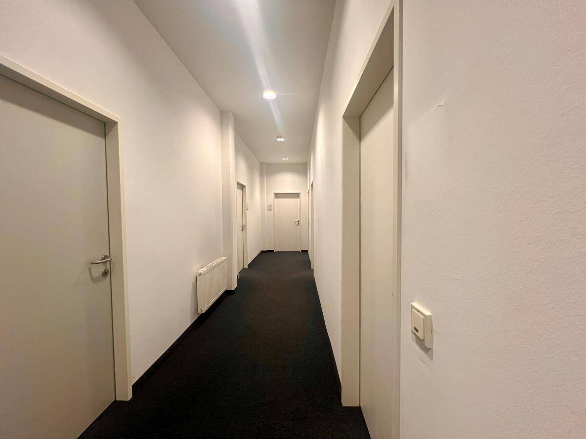 Langer, heller Flur mit mehreren Türen, die zu verschiedenen Büroräumen führen, und dunklem Teppichboden.