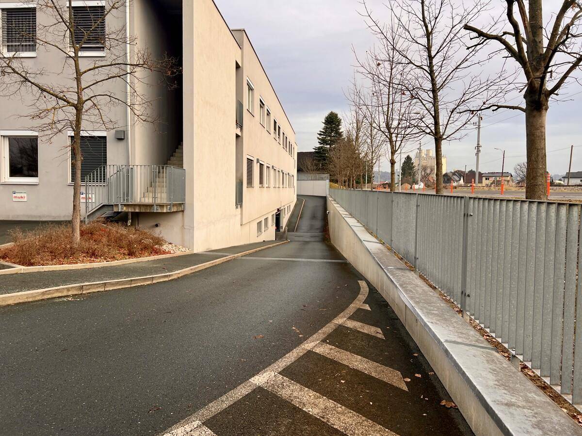 Zufahrt zur Tiefgarage eines modernen Bürogebäudes mit umliegenden Bäumen und einem Zaun.