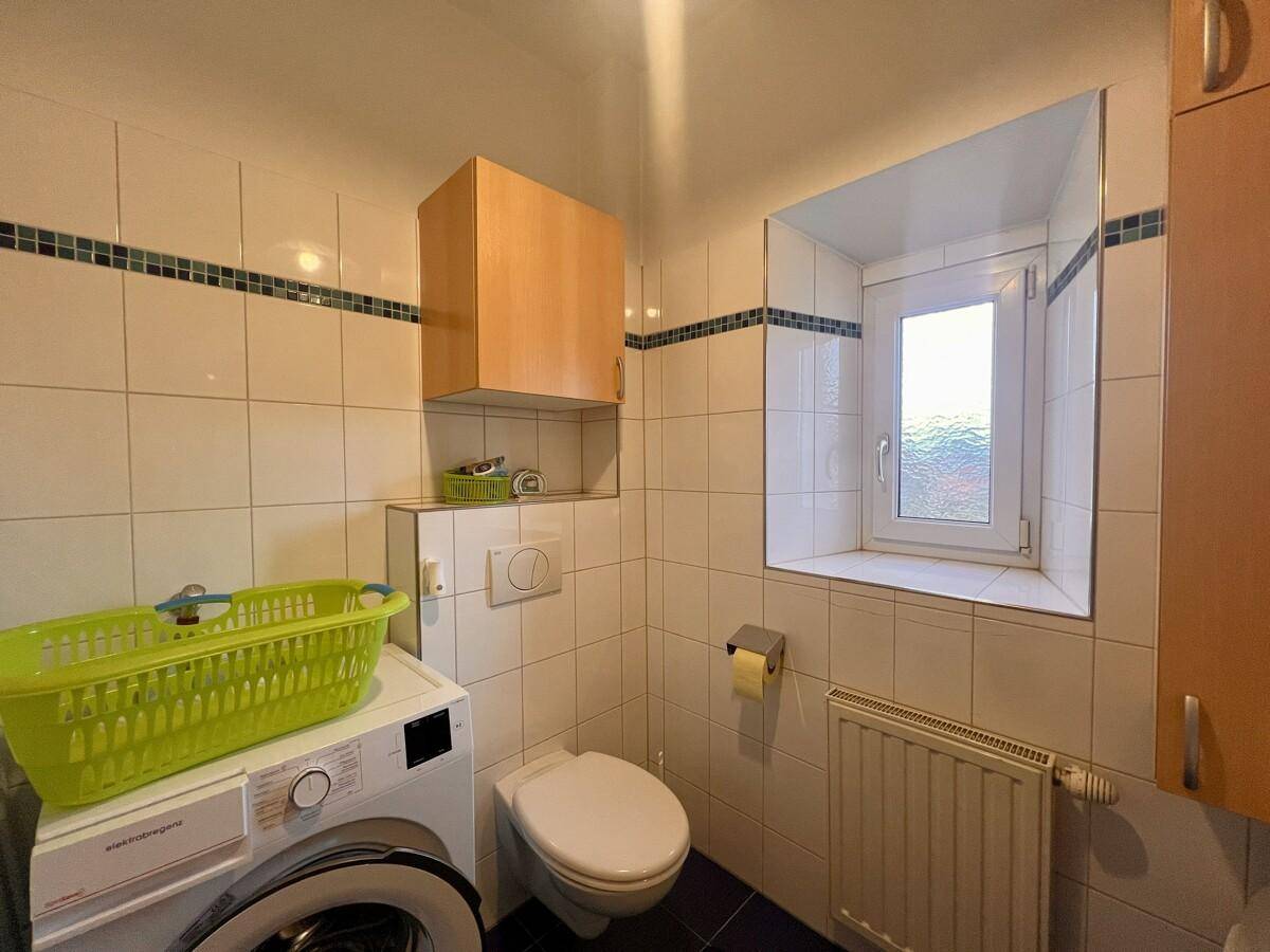 Kleines WC mit Fenster, Waschmaschine und Fliesenboden in praktischer Anordnung.