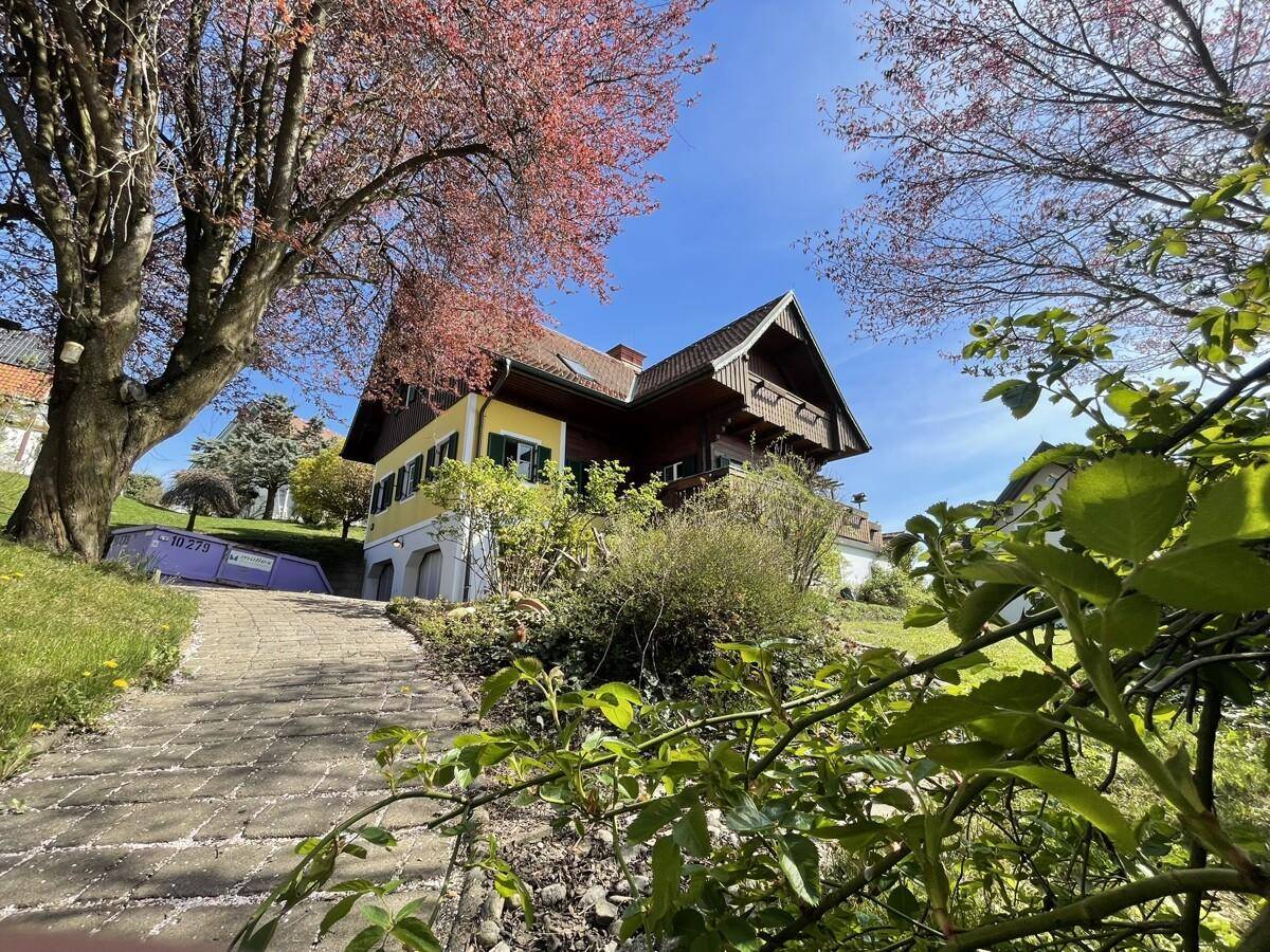 Hausansicht mit gepflastertem Weg, blühenden Bäumen und grüner Landschaft.