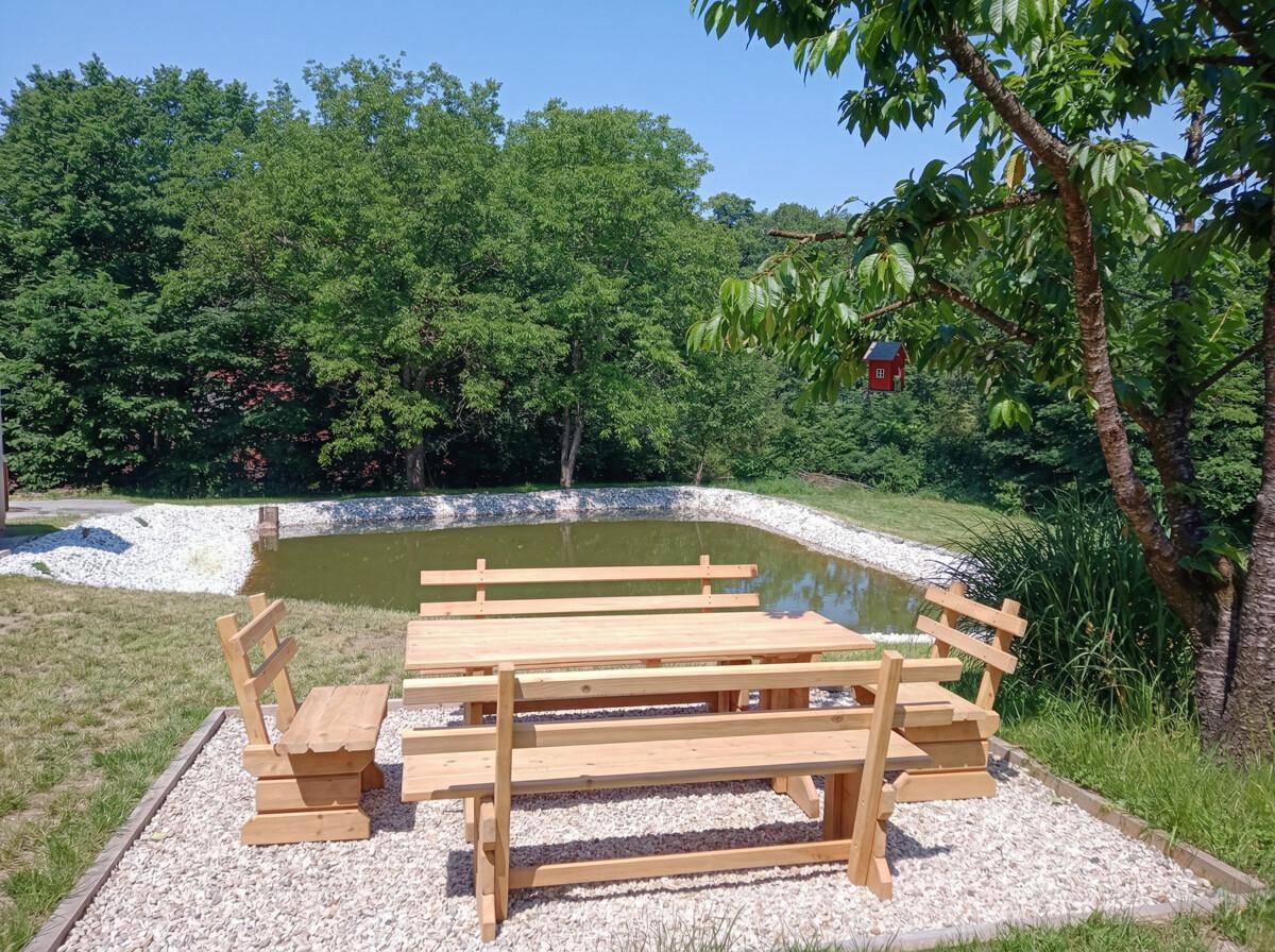 Gemütlicher Sitzbereich im Freien mit Holztisch und Bänken, Blick auf den Teich und den umliegenden Wald.