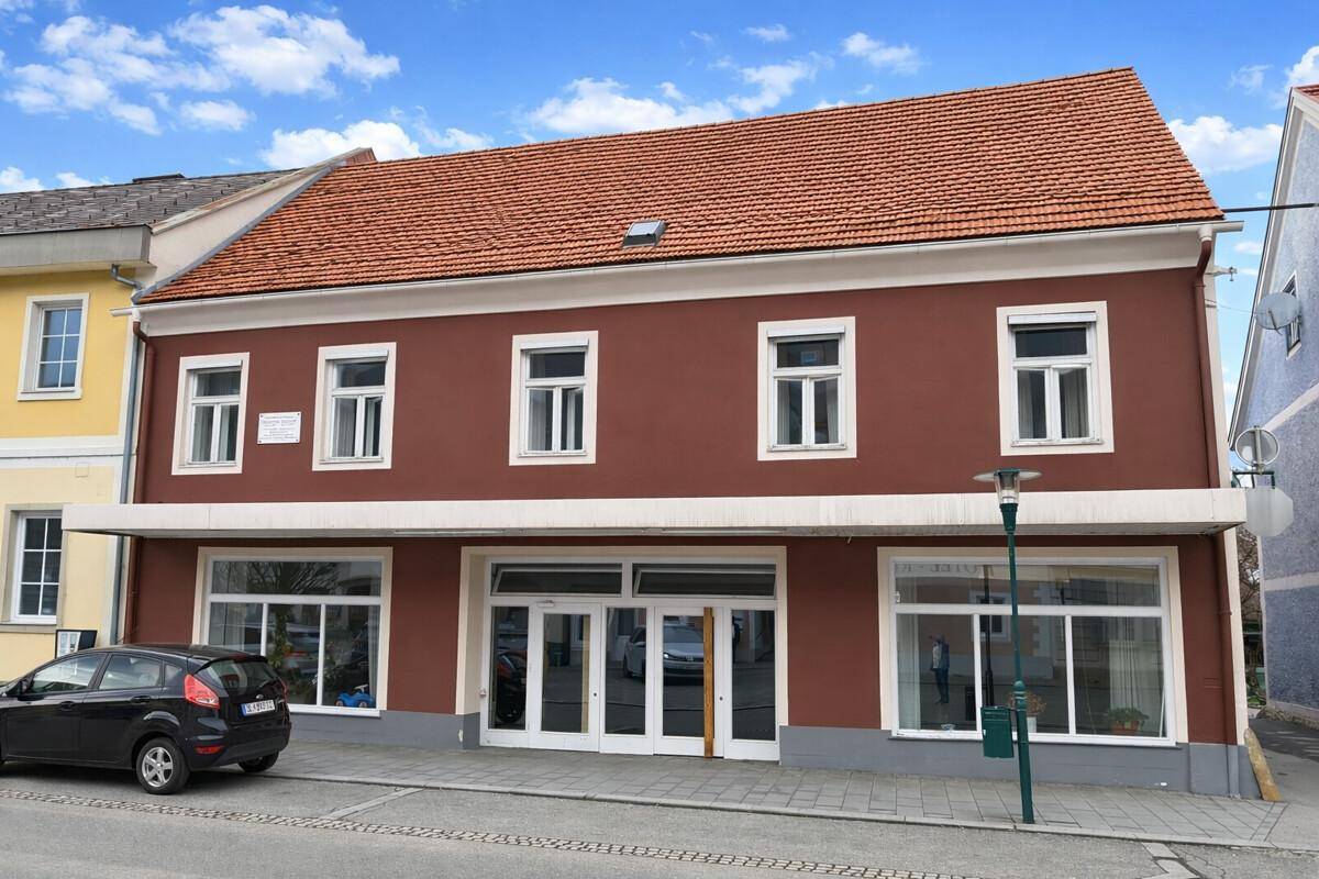 Vorderansicht des Gebäudes mit roter Fassade, großen Schaufenstern und traditionellem Ziegeldach an einer Straße.