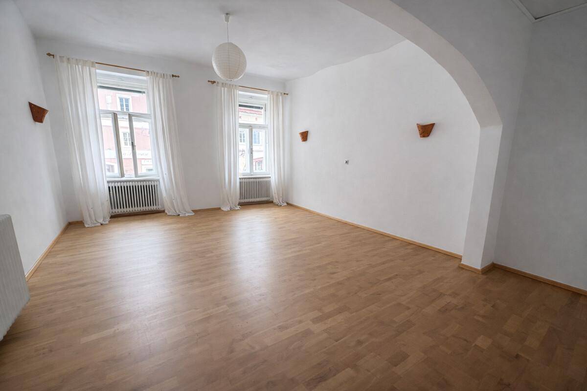 Geräumiges Wohnzimmer mit Parkettboden, hohen Decken und zwei großen Fenstern.