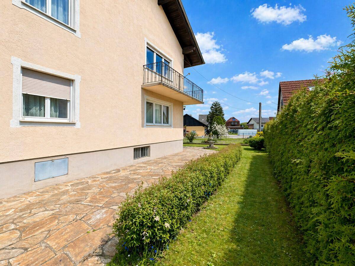 Seitenansicht des Hauses mit gepflastertem Weg, gepflegter Hecke und Rasenfläche unter blauem Himmel.