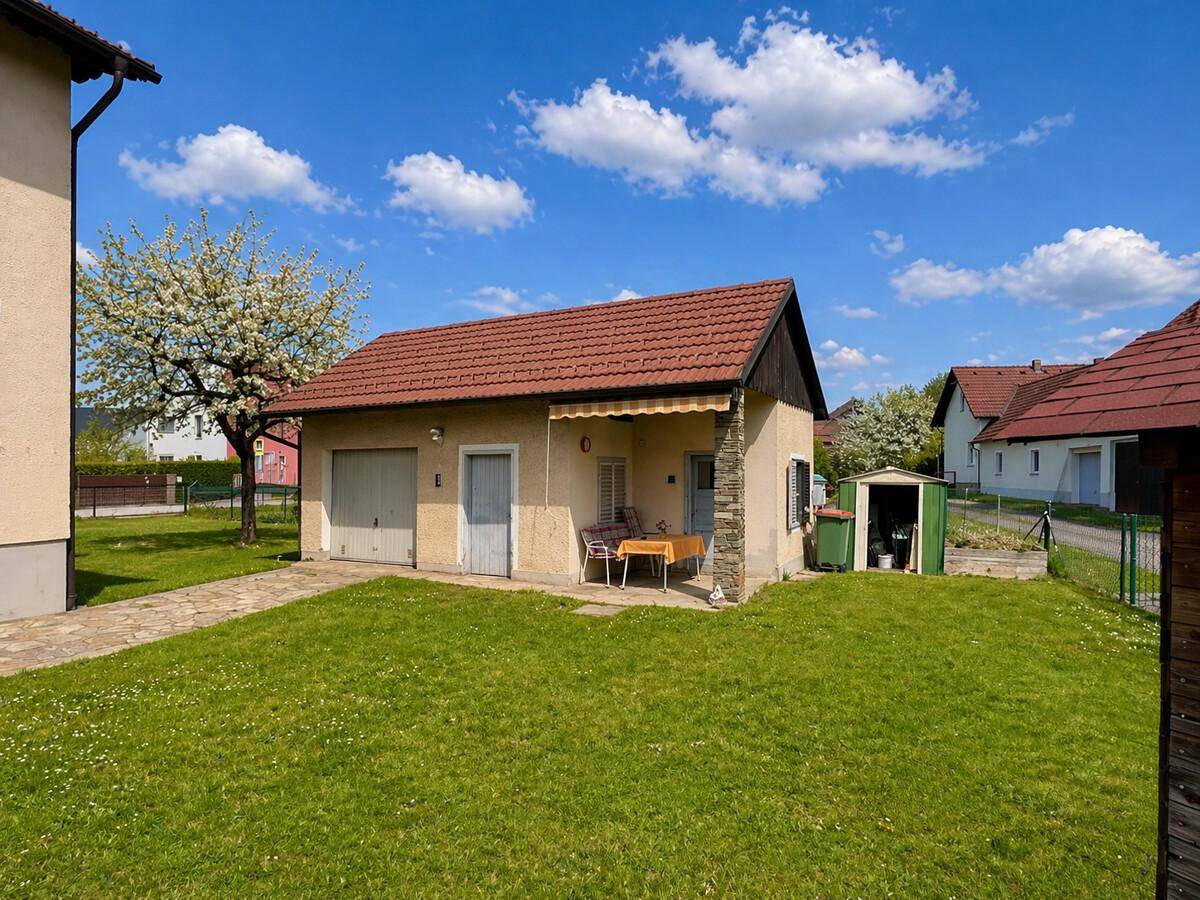 Ansicht des Nebengebäudes mit Garage, kleiner Terrasse und angrenzender Rasenfläche unter blauem Himmel.