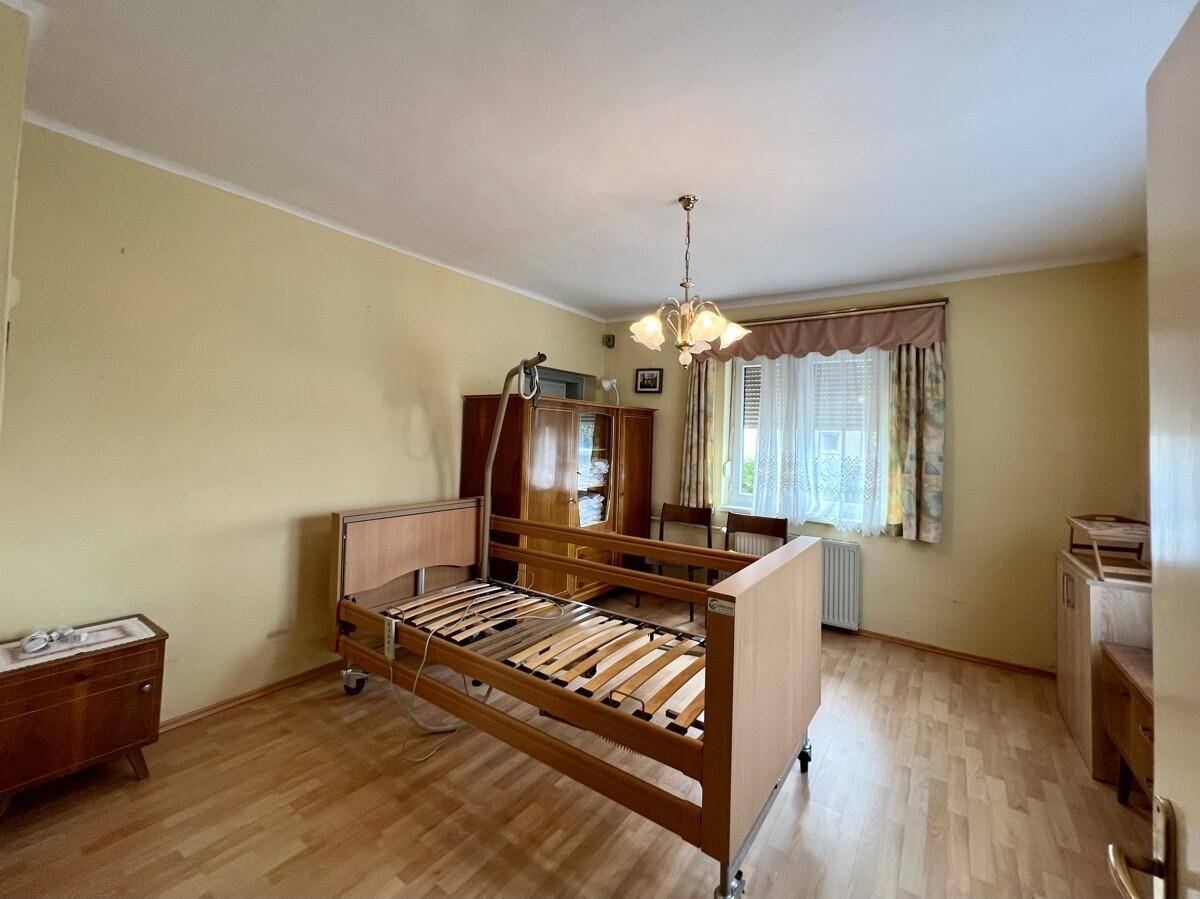 Schlafzimmer mit Pflegebett, Holzschrank und Fenster, das viel Tageslicht hereinlässt.