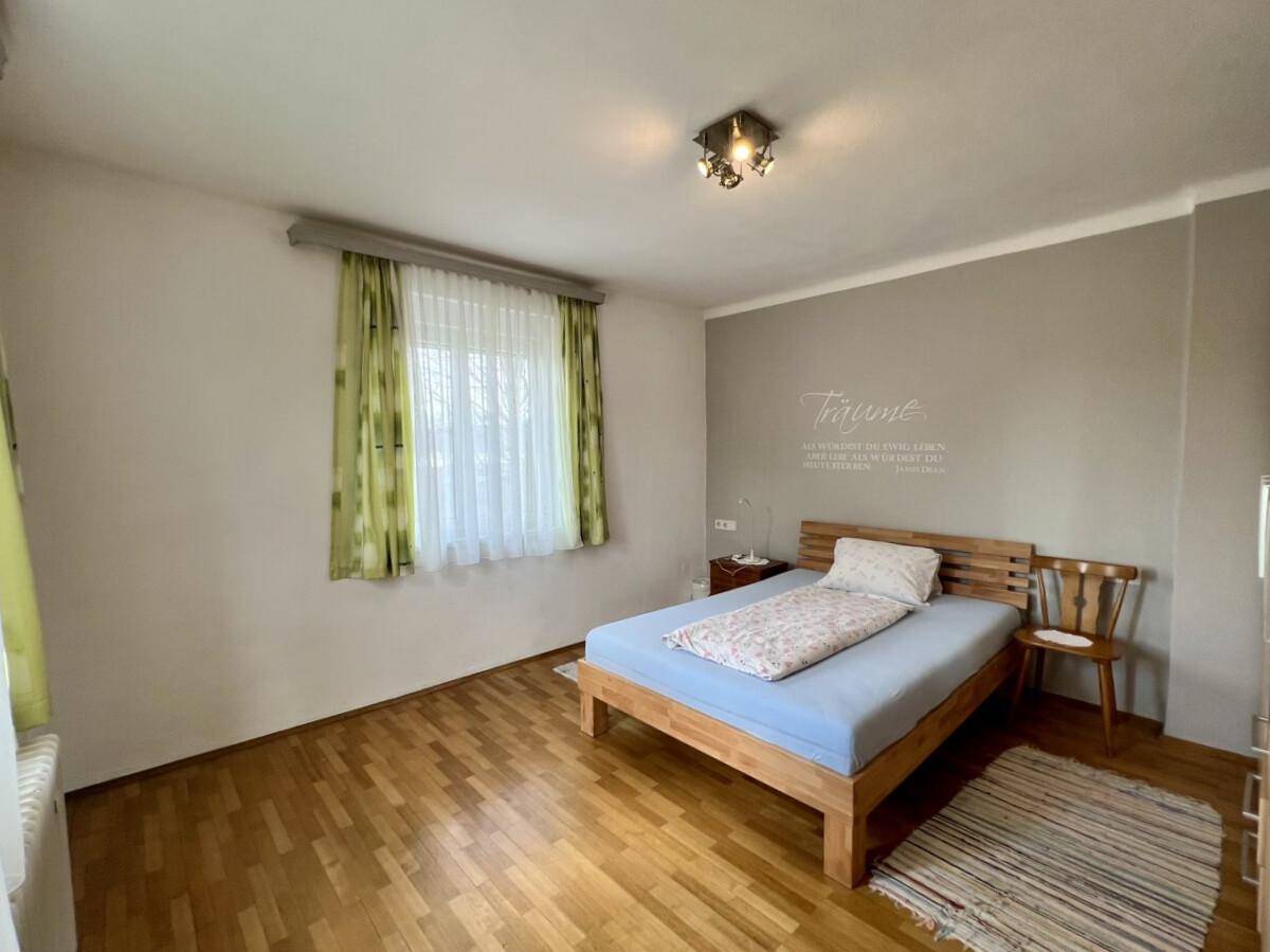 Helles Schlafzimmer mit Parkettboden, Holzrahmenbett und Fenster mit Vorhängen.