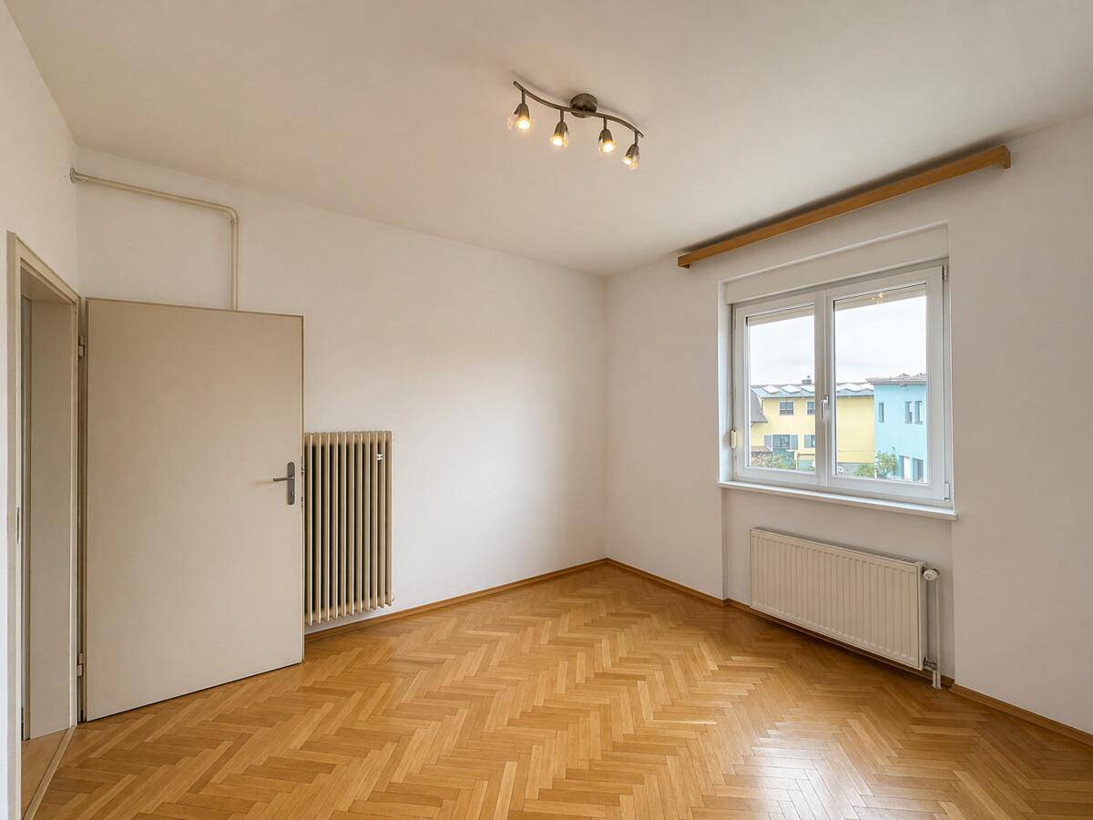Leeres Schlafzimmer mit Parkettboden, Fenster und Heizkörper, bereit zur individuellen Einrichtung.