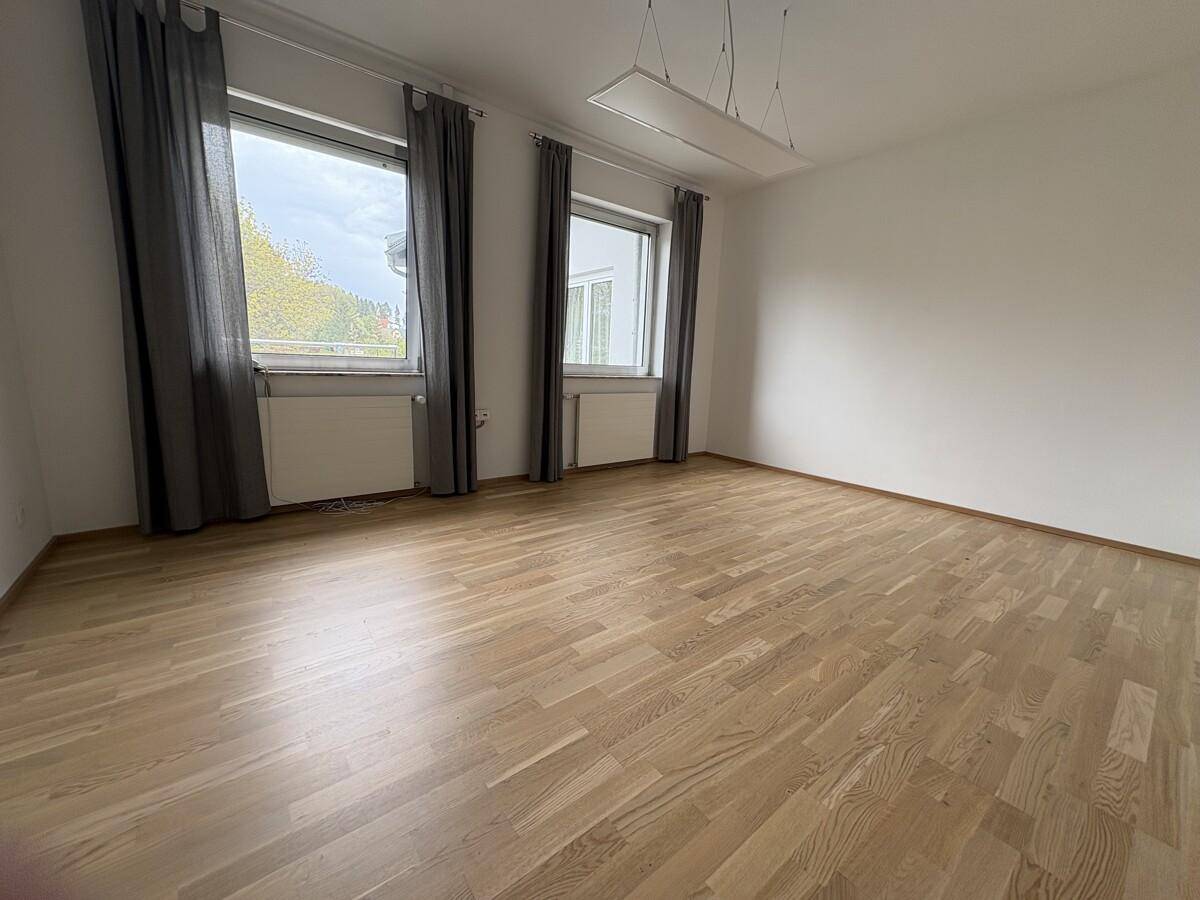 Großer, leerer Raum mit Holzboden und zwei Fenstern, bietet viel Tageslicht.