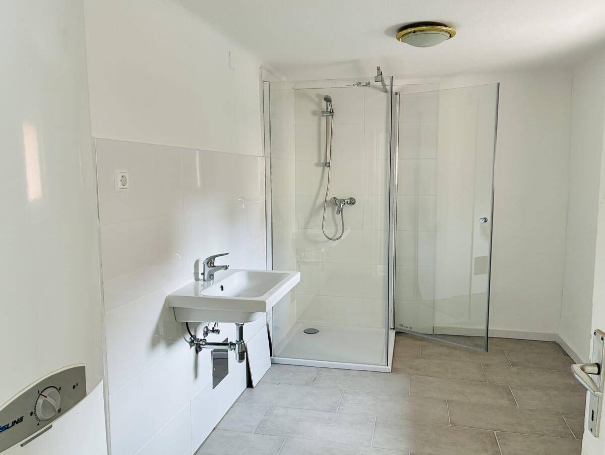 Badezimmer mit Duschkabine, Waschbecken und hellen Fliesen, inklusive eines Warmwasserbereiters.