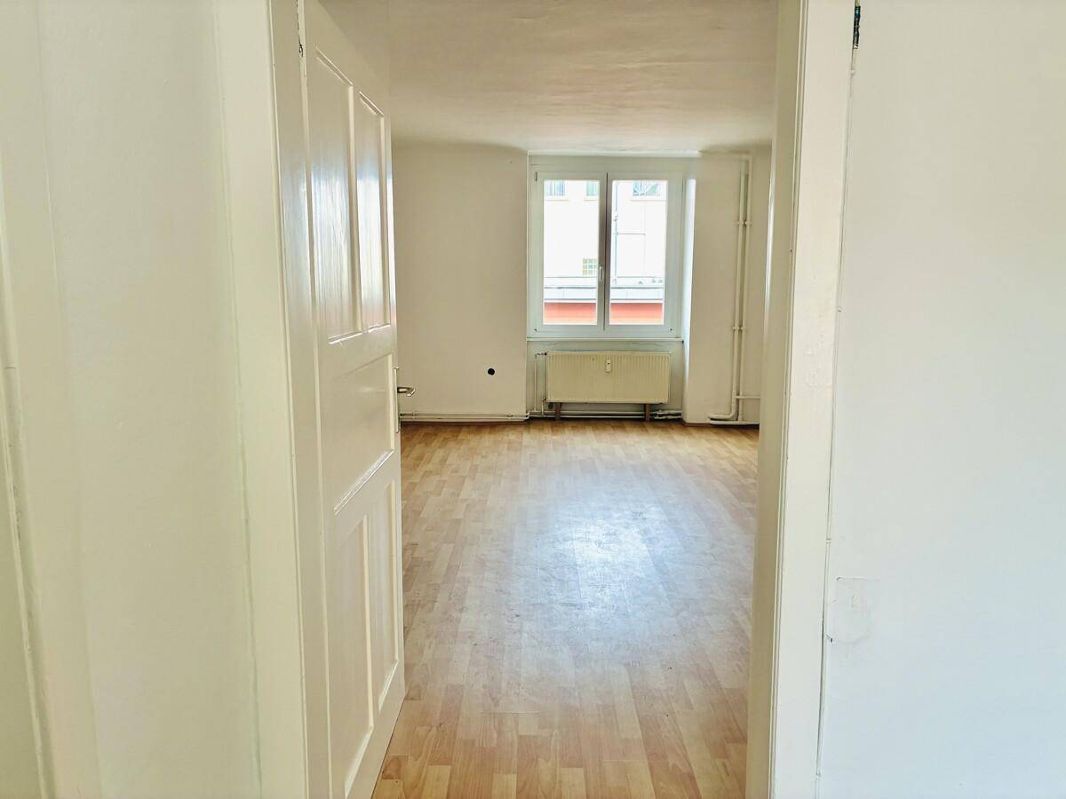 Geräumiges Wohnzimmer mit hellem Parkettboden, weißen Wänden und großem Fenster.