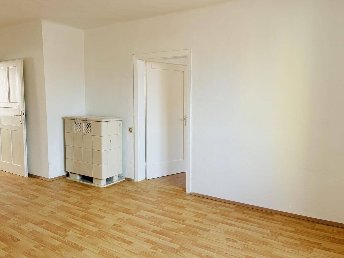 Wohnzimmer mit hellem Parkettboden, weiß gestrichenen Wänden und einem traditionellen Kachelofen.