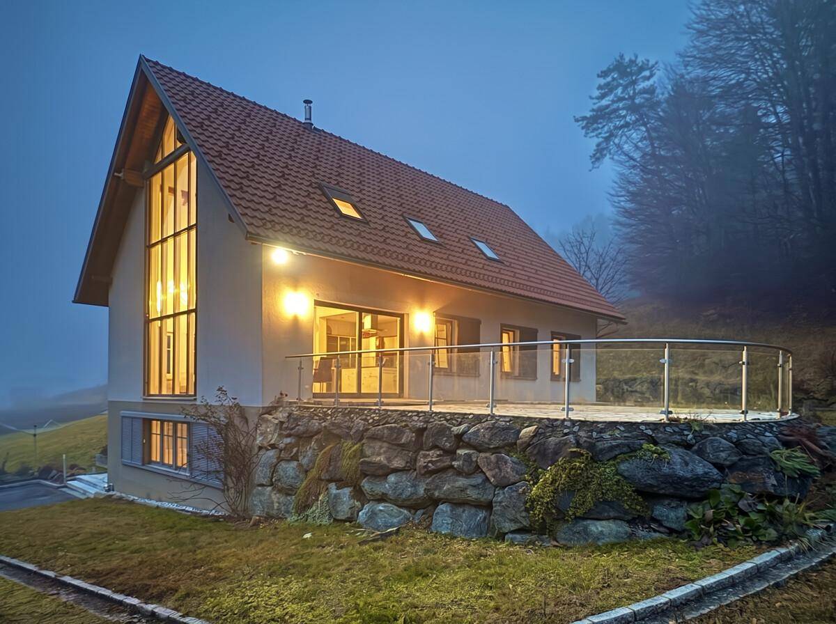 Modernes Haus in der Dämmerung mit beleuchteter Fassade und Terrasse, umgeben von Natur.