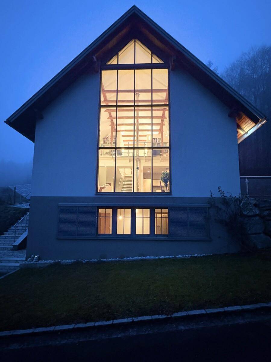 Moderne Hausfassade bei Nacht mit beleuchteten Innenräumen und großen Fensterfronten.