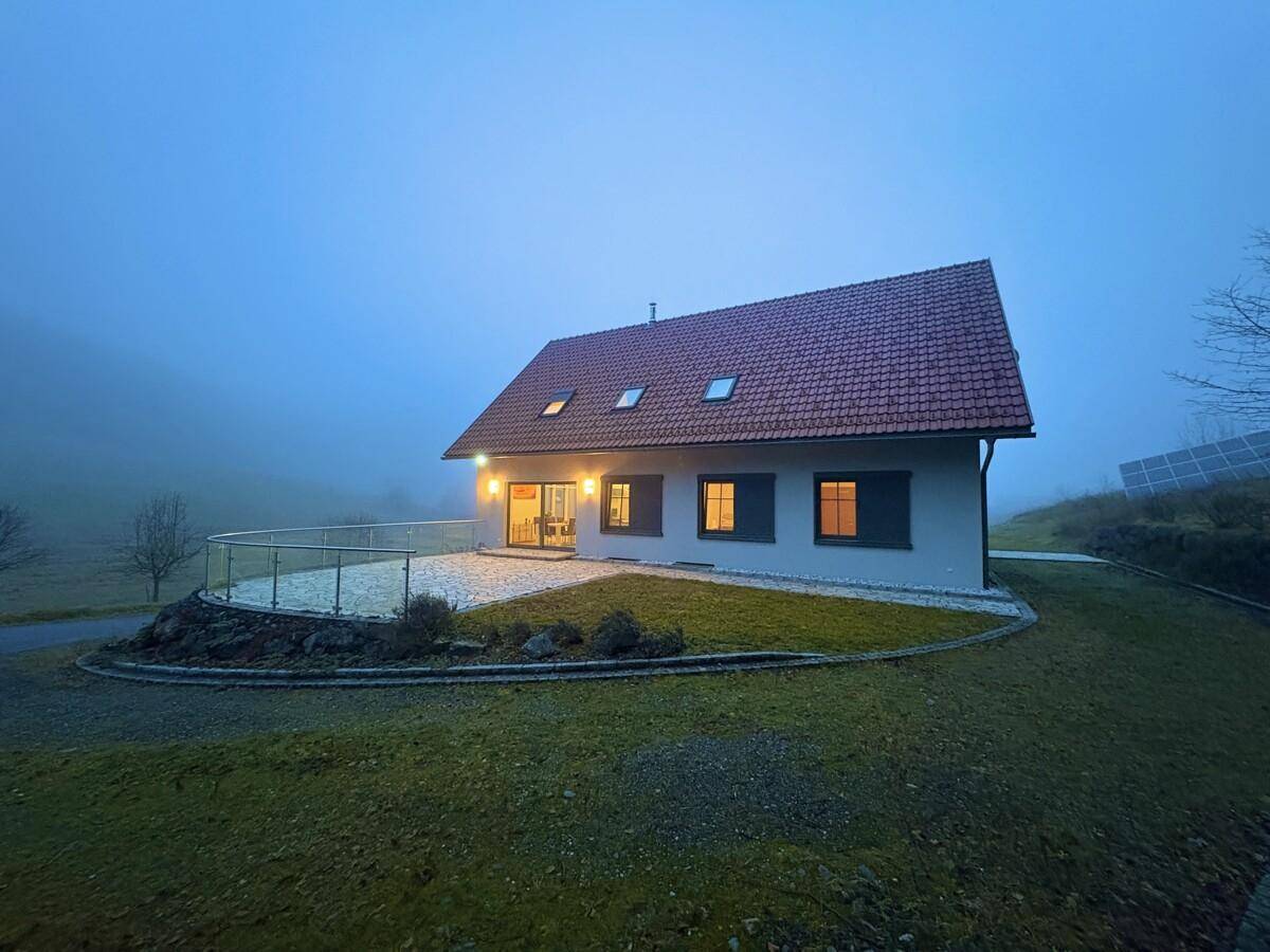 Haus in der Dämmerung mit beleuchteter Terrasse und Garten, umgeben von nebliger Landschaft.
