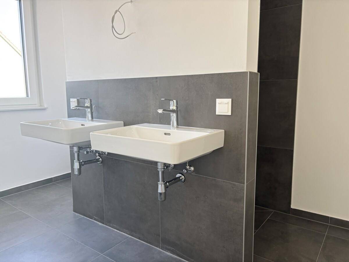 Modernes Badezimmer mit zwei Waschbecken, grauen Fliesen und Fenster für Tageslicht.
