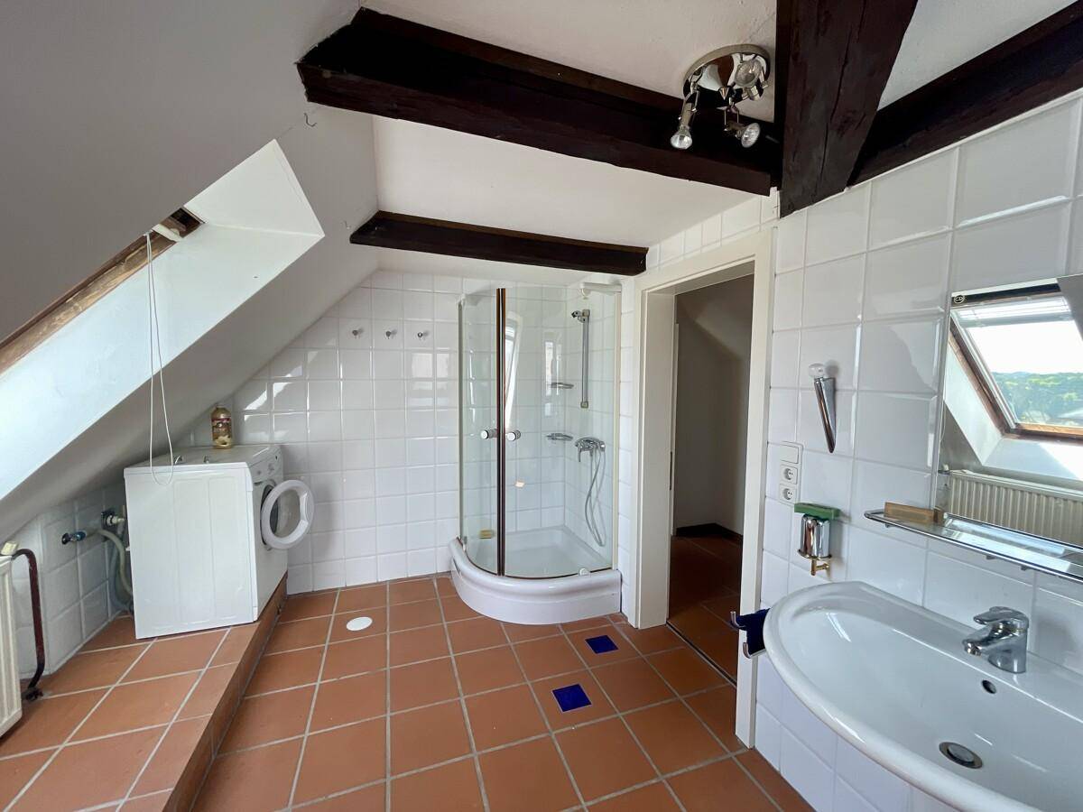Dachgeschossbad mit weißen Fliesen, Duschkabine und Waschbecken unter einer Dachschräge mit Holzbalken.
