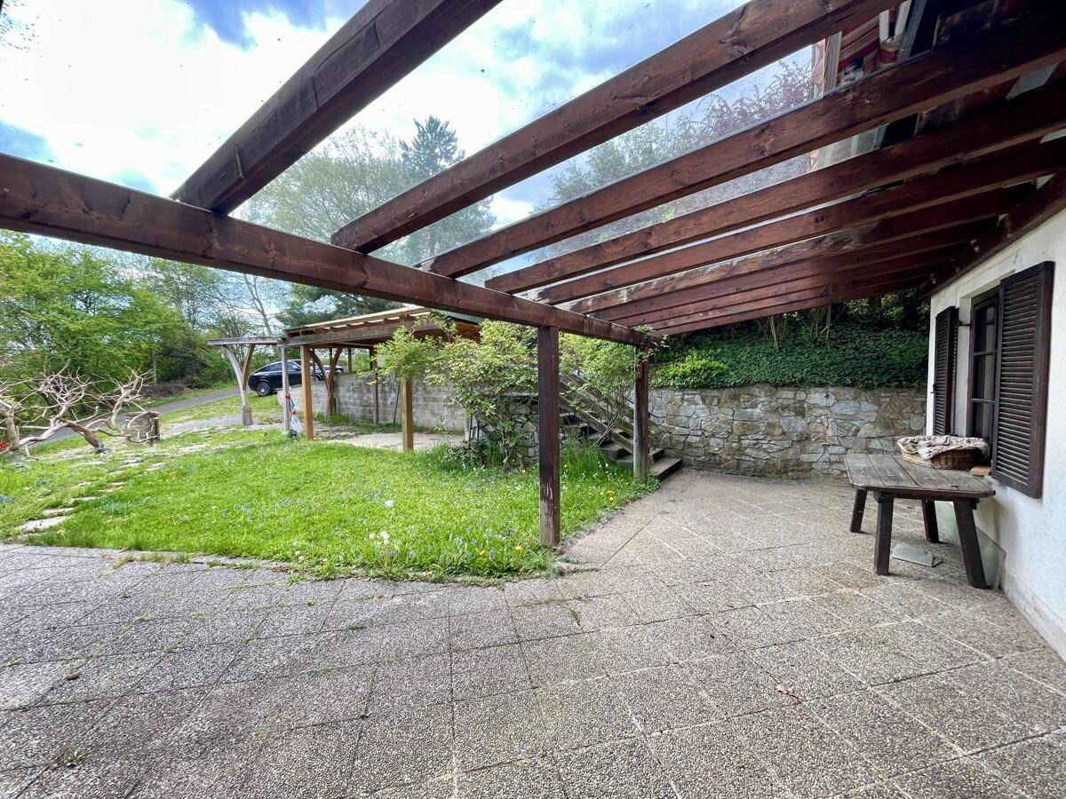 Gepflasterte Terrasse mit einer Holzpergola und einem kleinen Holztisch, angrenzend an einen Rasenbereich.