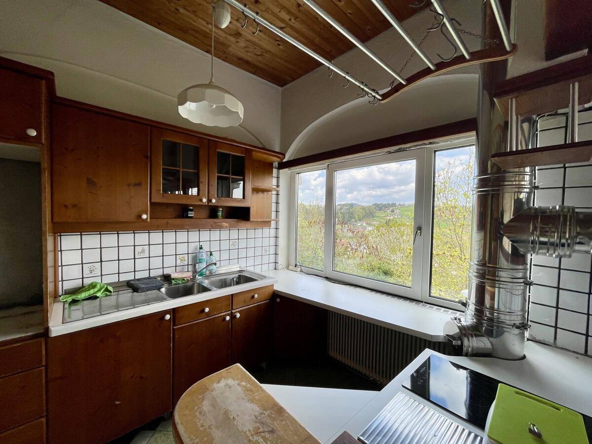 Traditionelle Küche mit Holzschränken, weißen Fliesen und einem großen Fenster mit Blick ins Grüne.