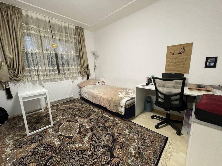 Schlafzimmer mit Einzelbett, Schreibtisch und Fenster mit hellen Vorhängen.