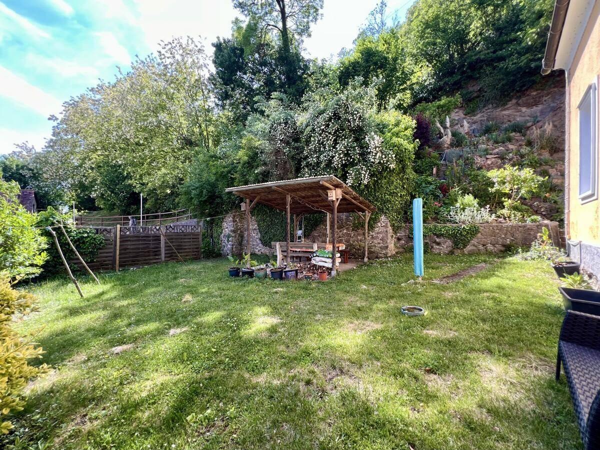Großer Garten mit Rasenfläche, überdachtem Sitzbereich und bewachsenem Hang.
