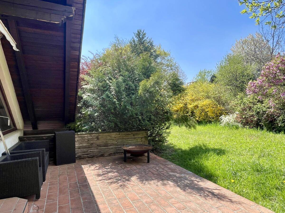 Sonniger Terrassenbereich mit Ziegelboden, Feuerstelle und angrenzendem grünen Garten.