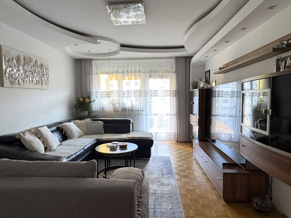 Gemütliches Wohnzimmer mit großem Sofa, Fernsehmöbeln und Parkettboden, hell durch Fenster.