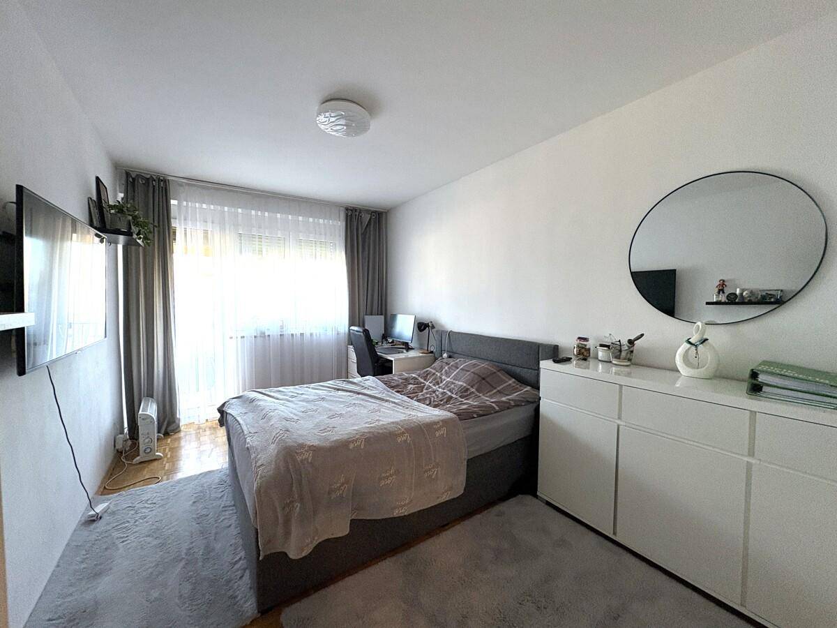 Helles Schlafzimmer mit Bett, Schreibtisch und weißer Kommode, gut beleuchtet durch ein Fenster.