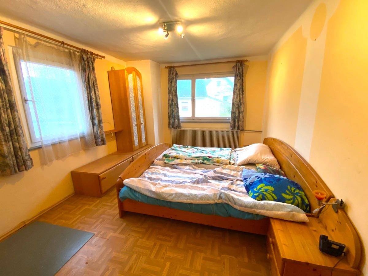 Schlafzimmer mit zwei Fenstern, Parkettboden und Holzmöbeln in heller Farbgebung.