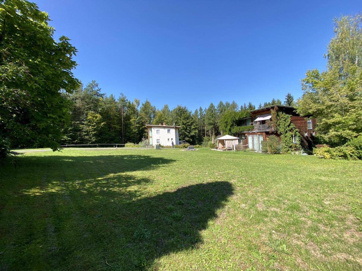 Weitläufige grüne Rasenfläche mit zwei Häusern und dichtem Baumbestand im Hintergrund unter blauem Himmel.