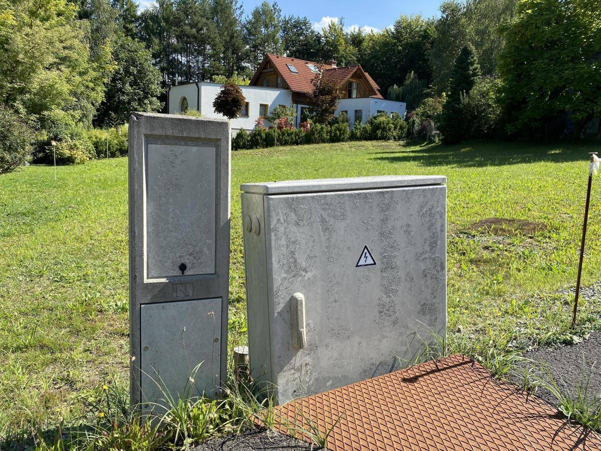 Zwei Versorgungsboxen auf einer grünen Wiese mit einem Haus und Bäumen im Hintergrund.