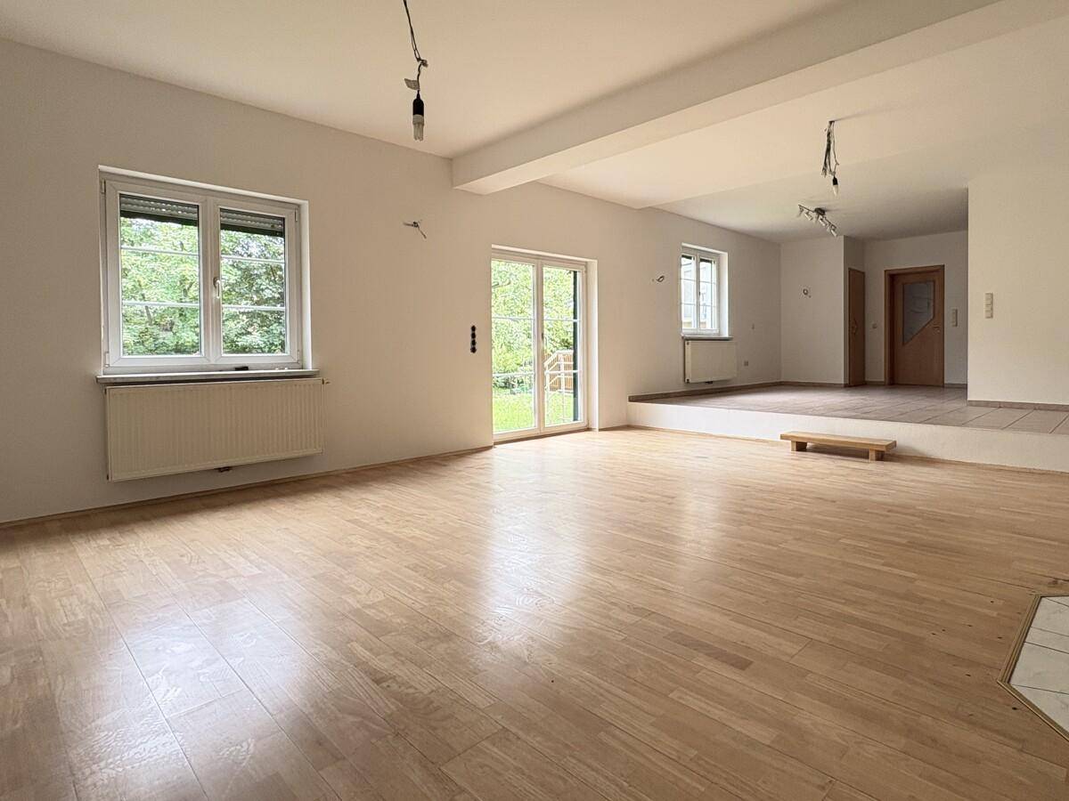 Heller Wohnbereich mit Parkettboden, großen Fenstern und Übergang zu einem erhöhten Bereich.